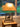 A wooden side table with a rustic lamp featuring a tan shade and a metal base with a moose and tree silhouette design. The table is placed near a window with a view of a green backyard and outdoor furniture.