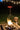 A nighttime outdoor scene featuring a fire pit with burning logs and glowing embers, surrounded by a brick border. A Muskoka electric patio heater with red glowing elements is mounted on a tripod stand nearby. String lights with warm bulbs hang above, illuminating the grassy area, with a tree stump and marshmallow roasting sticks visible in the background.